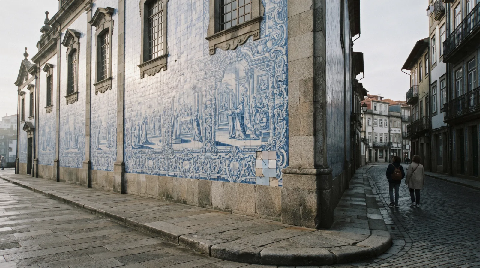 The blue-and-white tiled side wall of the Igreja do Carmo in morning light, one of the Porto churches, two visitors small and seen from behind on the pavement below